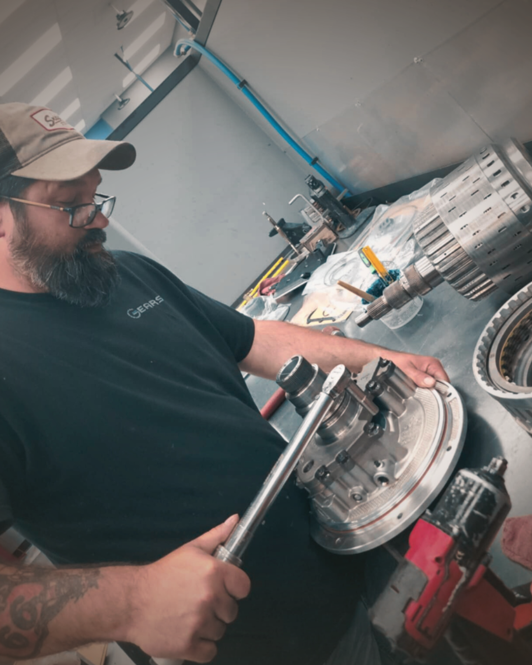 A bearded man wearing a black t-shirt and a baseball cap uses a socket wrench to tighten a silver metal clutch plate on a workbench in an automotive workshop.